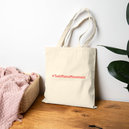 Beige tote bag with '#TaskMamaMovement' text on a wooden surface with a plant in the background