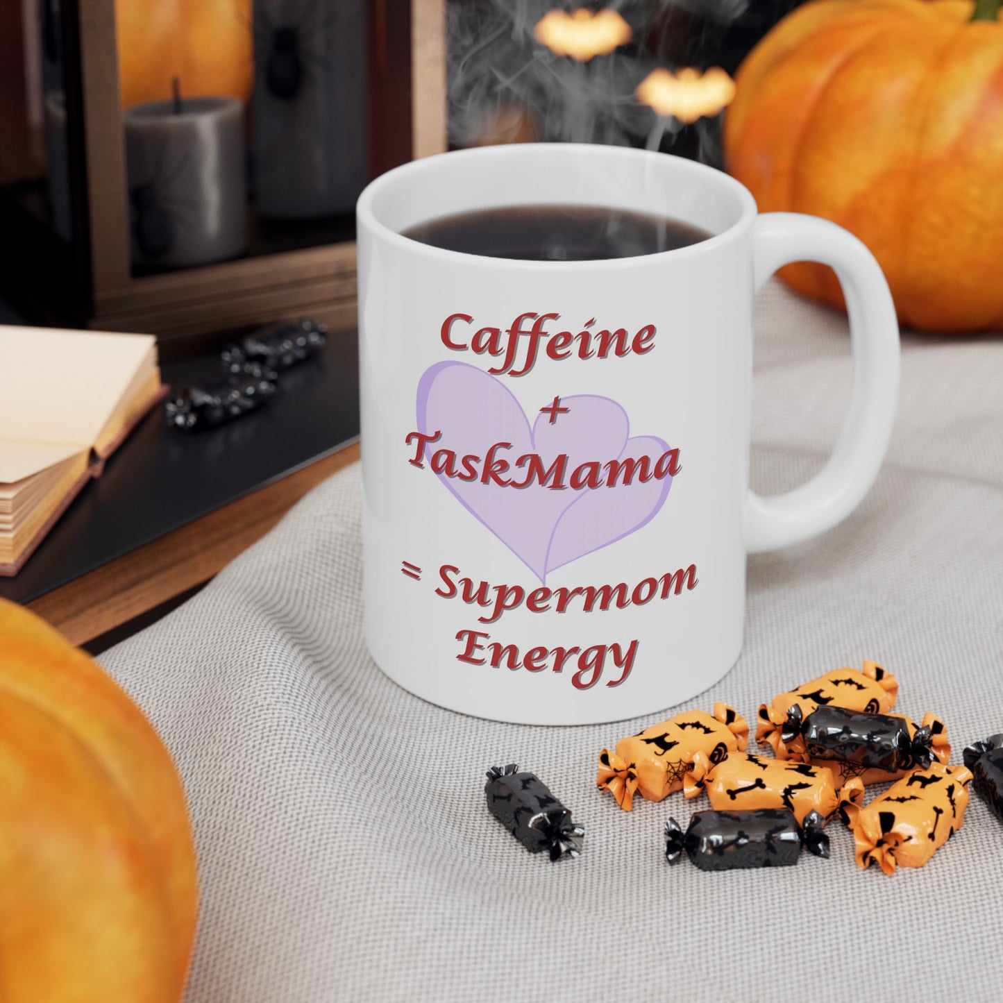 White mug with 'Caffeine + TaskMama = Supermom Energy' text, surrounded by Halloween-themed treats and pumpkins.