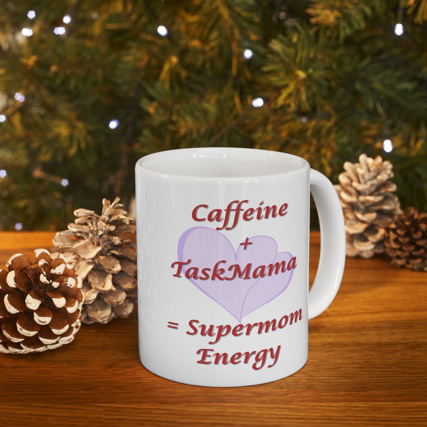 White mug with text 'Caffeine + TaskMama = Supermom Energy' on a wooden surface with pinecones and a Christmas tree in the background.