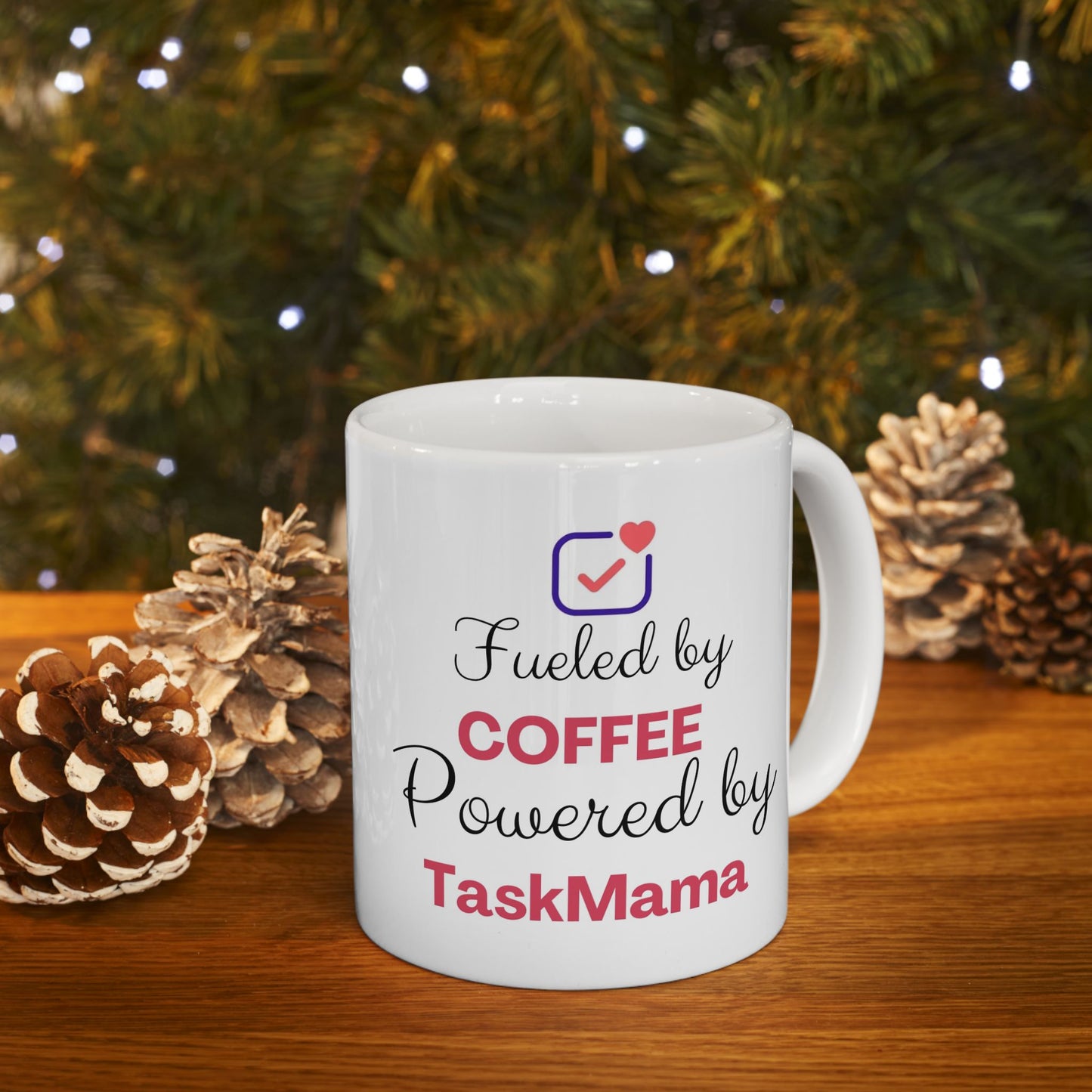 White mug with text 'Fueled by Coffee Powered by TaskMama' in front of a Christmas tree and pinecones.